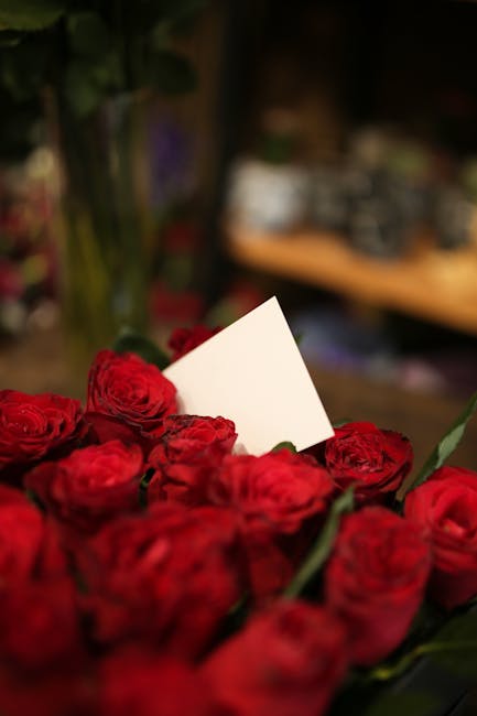 A close-up view of a vibrant bouquet of fresh red roses with lush petals and green stems, arranged densely together. The bouquet is partially wrapped in clear cellophane, with a white card inserted am