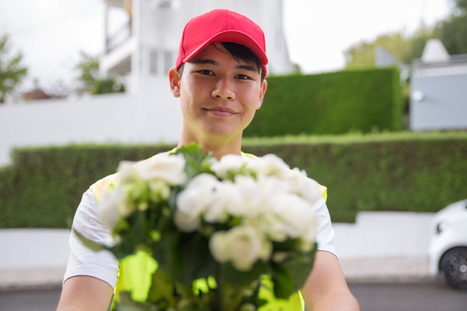 A delivery person in a red cap and high-visibility vest is smiling while holding a bouquet of fresh white flowers, possibly roses or peonies, wrapped in clear cellophane. The bouquet features lush, vi