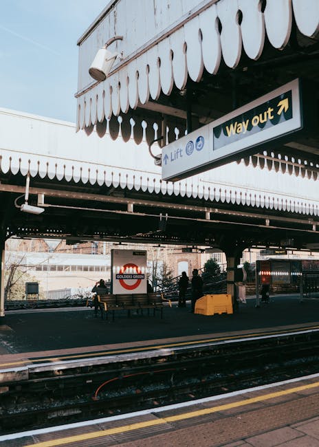The image depicts a floral arrangement held by a person at Golders Green underground station, featuring a bouquet of fresh flowers with pastel pink roses, white lilies, and purple irises, wrapped in l