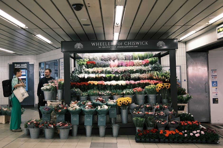 A floral display of fresh flowers arranged in a tiered setup within a busy underground station, featuring various types such as roses, sunflowers, tulips, and chrysanthemums in vibrant colors includin