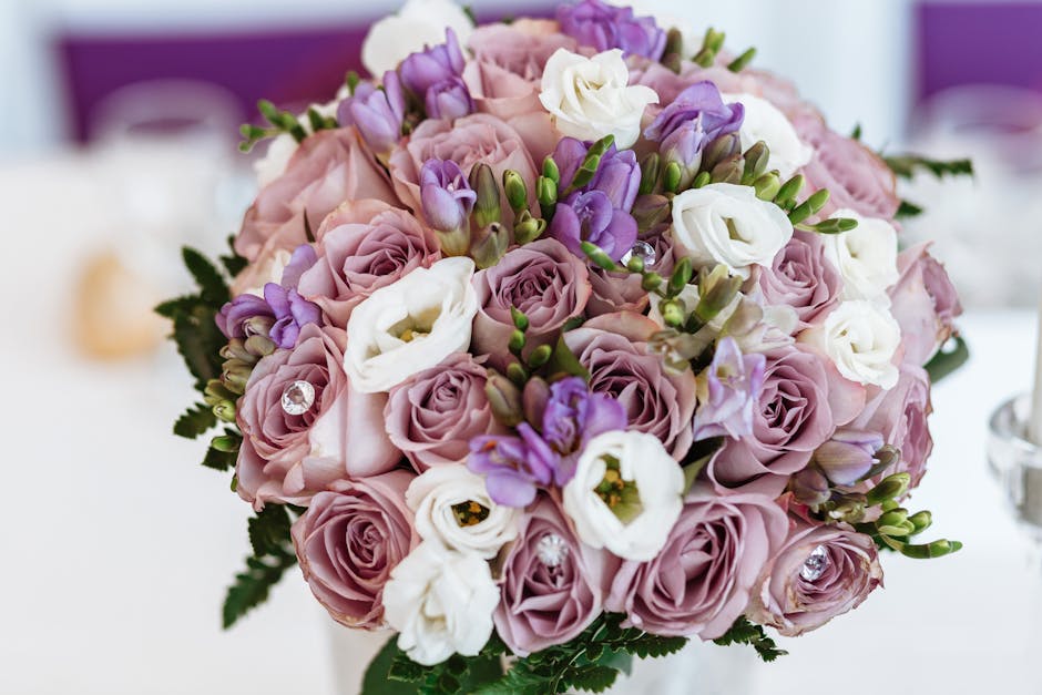 A close-up of a floral bouquet featuring soft pink roses, white lisianthus, and purple freesia arranged in a compact, rounded style. The flowers are fresh with open petals and sturdy green stems, acce